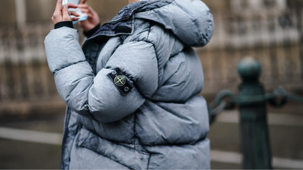 Человек в серой куртке Stone Island с логотипом бренда на рукаве снаружи.